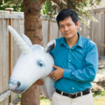 Vu Le poses outside in front of a tree and fence with a blow-up unicorn head